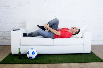 Rolgordijnen fanatic football fan lying on couch sofa with ball on green grass carpet emulating soccer stadium pitch mocking player in pain hurt on ankle © Wordley Calvo Stock