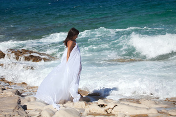 Fototapeta na wymiar Woman in white cape dress enjoys on the rocky beach on the sea