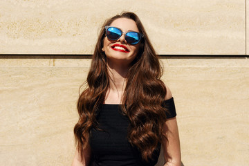 Closeup portrait of young happy smiling beautiful girl with long brunette curly hair posing against wall and smiling on a sunny warm day