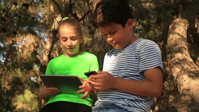 Happy young boy and girl sitting on the branches of tree with digital tablet and smart phone