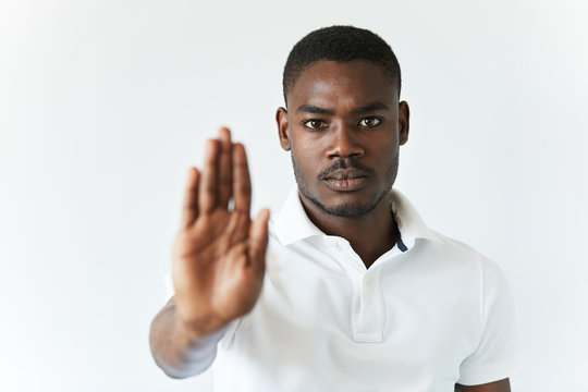 Headshot of dark-skinned young man wearing white polo shirt, preventing you from doing something with his open palm, looking at the camera with serious expression. Selective focus on the face