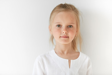 Little European girl in white children clothes looking peacefully at the camera indoors. Calm child standing quietly in restful manner with angelic look and innocent appearance.
