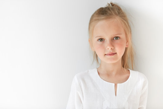 Blond Caucasian female child with green eyes standing quietly indoors and looking straight forward in good and sunny day. White colors make little girl look like a little angel or innocent baby.