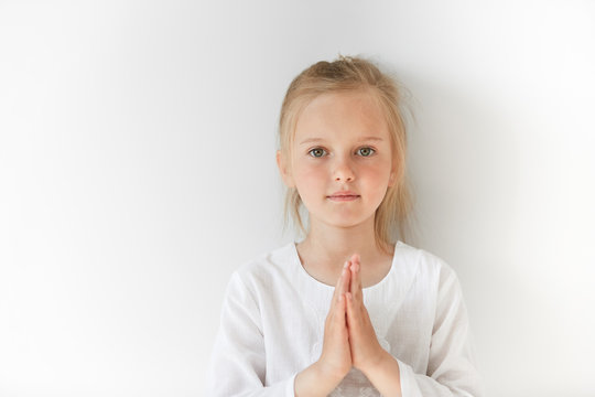 Little European girl in white clothes praying and looking forward with calm and pretty facial expression. Blond child with green eyes watching you peacefully in morning light.