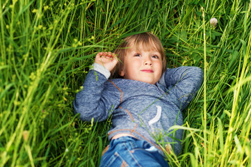 Candid portrait of adorable little boy of 4-5 years old, wearing blue hoody, playing alone outdoors, laying on grass