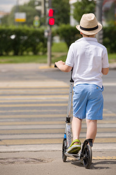 Cute Kid Boy In Straw Hat On A Scooterstanding On Crossroads And Waiting For The Light To Turn Green. Child Waiting On Green Light On The Road. Schoolboy On Scooter Crossing The Road On Sunny Day.