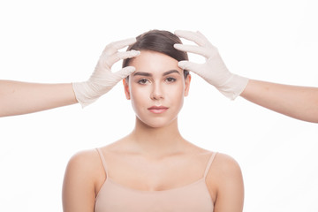 Fototapeta premium Handmade beauty. Beautiful young woman keeping eyes opened while two hands in medical gloves touching her face isolated on white background.