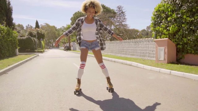 Slow motion view of young afro-american woman on vintage roller skates rides through the upmarket village streets and showing her driving skills.