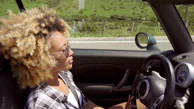 Young woman with a trendy afro hairstyle driving her sports car or cabriolet along a rural highway  high angle close up side view