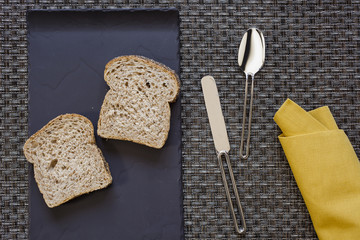 Black flat plate with two slices of bread and mustard yellow folded napkin spoon and knife aside on woven grey thick wire warp textured background