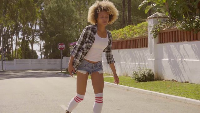 Young woman dressed in plaid men's shirt  white top  shorts and knee-high socks rides through the upmarket village streets towards the camera in slow motion.