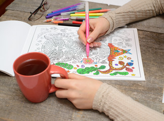 Fototapeta na wymiar Woman coloring an adult coloring book and drinking tea, new stress reliving tool