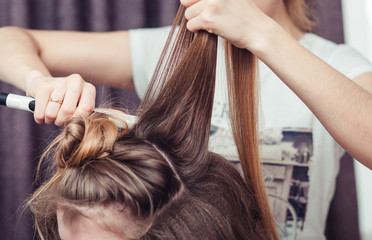 Hairstylist using a curling iron of a female client in a hairdressing salon, closeup view