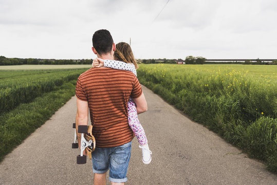 Father with skateboard carrying daughter on country lane