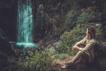 Beautiful woman hiker sitting near waterfall in deep forest.