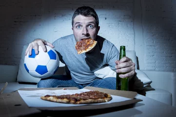 Tuinposter football fan man watching soccer game on tv at home sofa couch with soccer ball and pizza in his mouth © Wordley Calvo Stock