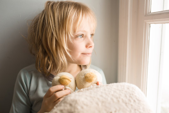 Young boy holding soft toys, sitting beside window, looking at view