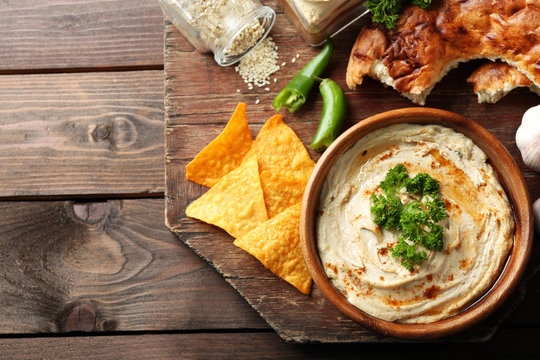 Wooden bowl of tasty hummus with chips and parsley on table