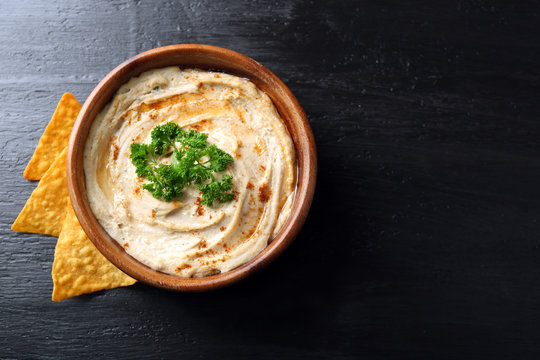 Wooden bowl of tasty hummus with chips and parsley on table