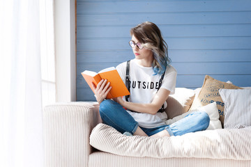 Young smart female student studying diligently at home near large window. Balcony, sofa, soft pillows. Orange textbook. Her hair is blue, she is dressed in jeans and a white T-shirt.