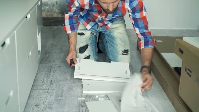 Troubled man with instruction trying to match pieces of furniture on floor at home
