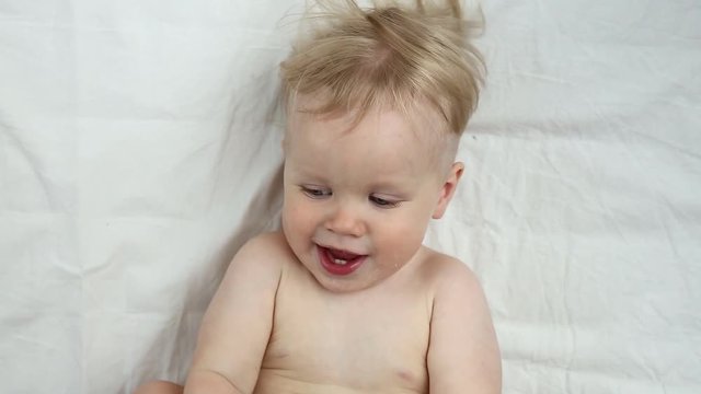 Adorable little baby is looking into the camera and is happy on a white bed sheet. The baby looks around and then smiles and laughs. Baby boy with blue eyes smiles at home