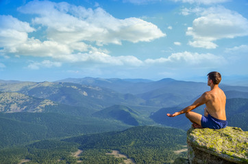 Naklejka na ściany i meble on top of a high mountain cliff in front of a man sitting in the lotus position and meditates. a man sitting on top of a mountain in the lotus position.