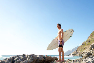 Attractive young man standing on beach carrying surfboard