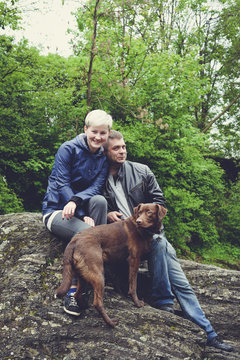 Happy Family Couple With Dog Hiking Traveling In The Forest. Beautiful Sporty Woman And Young Handsome Man In Leather Jacket And Cute Pedigree Dog Together Outdoors In The Forest Mountains Near River