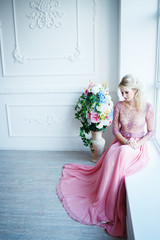 Beautiful blonde woman in evening pink dress posing in studio. beautiful model posing in a classic white interior.