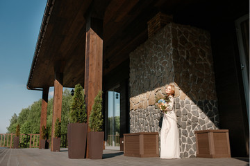 невеста с букетом в стиле бохо стоит прислонившись к декоративной стене из дикого камня на открытой веранде в солнечный день