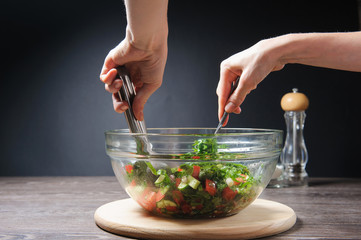 Cooking salad. Young woman cooking vegetable salad at home. Hands stirs vegetarian salad with fresh tomatoes, cucumber on wood table against dark background on rustic kitchen.