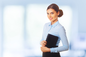 Portraitof happy smiling young cheerful businesswoman in blue clothing. Hold diary in her arms. Standing in her office, blue background.