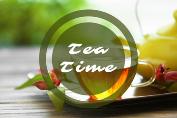Text Tea Time and glass cup of tea with teapot on wooden table against blurred background