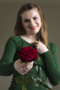 Eine junge braunhaarige Frau in grünem Pullover und mit rotem Lippenstift hält eine rote Rose in den Händen