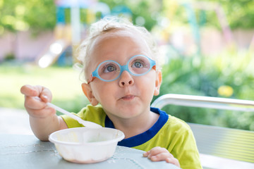 Ребенок в очках сидит за столом перед тарелкой  с ложкой на улице