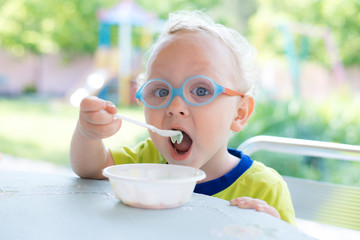 Ребенок в очках ест на природе мороженное ложечкой из тарелки 
