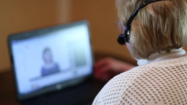 Woman with headphones with microphone talks to a customer via computer. Woman with pc communicates through skype. Woman with laptop. Senior woman with headphones and notebook speaks with customer