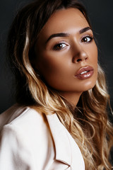 Beautiful, sexy blonde girl with bronze tan in a white coat and beige shoes posing in a studio on a dark background.