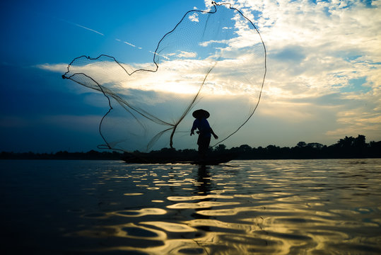 Silhouette of asian fisherman on wooden boat in action casting a net for catching freshwater fish in nature river in the early morning before sunrise