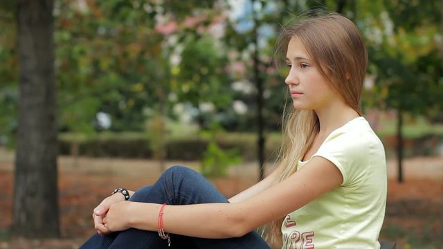 Sad pensive girl teen teenager sitting on park bench in early autumn