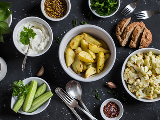 Baked cauliflower and potatoes, fresh cucumbers and herbs, sauces and spices on a dark stone background. Healthy vegetarian lunch or snack in a rustic style. Top view