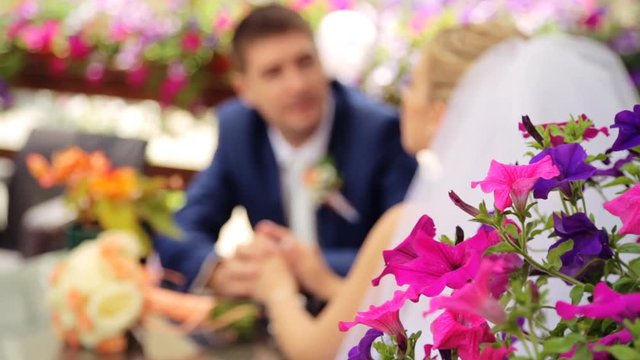 Handsome groom telling something to his beautiful bride sitting at a table in summer terrace with violet flowers on background