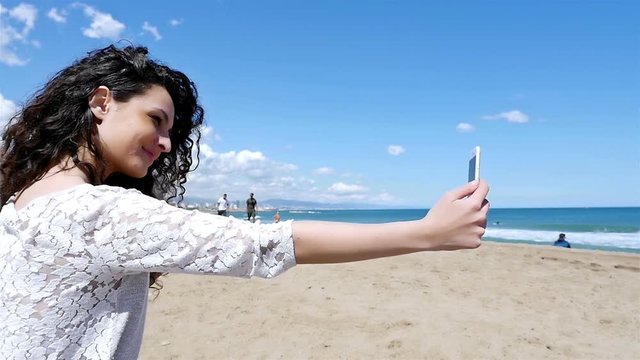 Portrait of pretty young woman taking selfie with a smart phone on the beach, slow motion