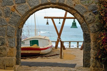 Fototapeta na wymiar View through an arch onto a boat and a wooden construction with green bells on it, in the background the ocean