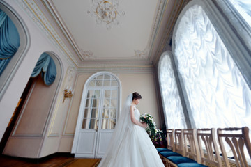 Obraz na płótnie Canvas Beauty bride in bridal gown with bouquet and lace veil indoors. Beautiful model girl in a white wedding dress. Female portrait of cute lady. Woman with hairstyle