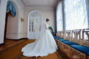 Fototapeta premium Beauty bride in bridal gown with bouquet and lace veil indoors. Beautiful model girl in a white wedding dress. Female portrait of cute lady. Woman with hairstyle