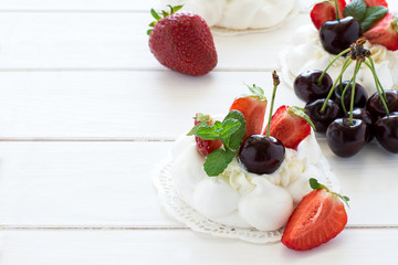 Homemade small pavlova meringue cakes with mascarpone cream, strawberries, cherries and fresh mint leaves on white wooden background