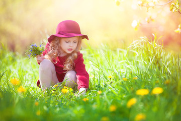 Beautiful little girl picking flowers in a meadow. Summer sunny day, lots of greenery and flowers.Blonde girl in burgundy dress.