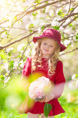 Beautiful little blonde girl in hat and burgundy dress. In her hand bouquet. Summer sunny day, lots of greenery and flowers